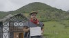 A male Agroecology course graduate smiles and holds up his certificate, whilst stood in front of a black and white building and lush green hillside. thumbnail